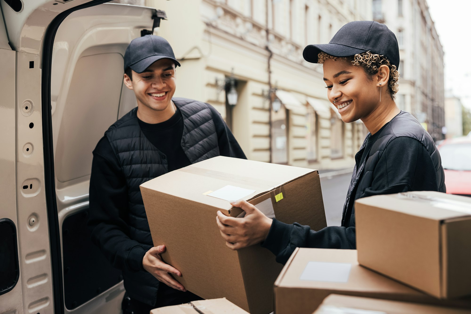 Two smiling couriers unloading cardboard boxes from van in the city